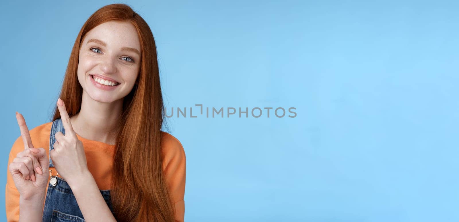 Lifestyle. Happy tender redhead girl sincerely smiling white teeth helpful look camera excited give hand pointing upper left corner introduce sale offer recommend try promo standing blue background.