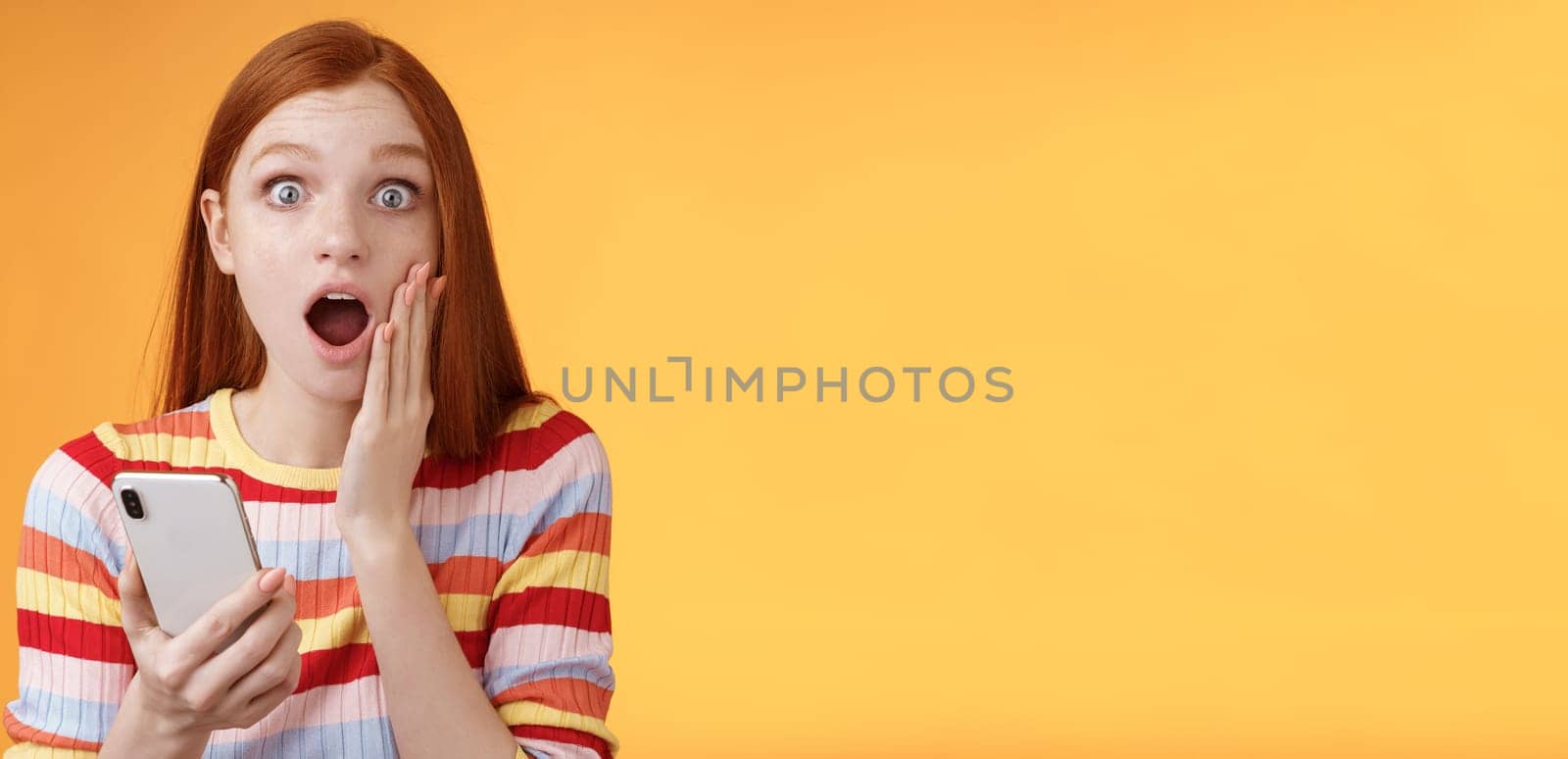 Concerned shocked emotive redhead girl find out stunning news look camera drop jaw gasping impressed touch cheek hold smartphone finish reading impressive disturbing story, orange background.