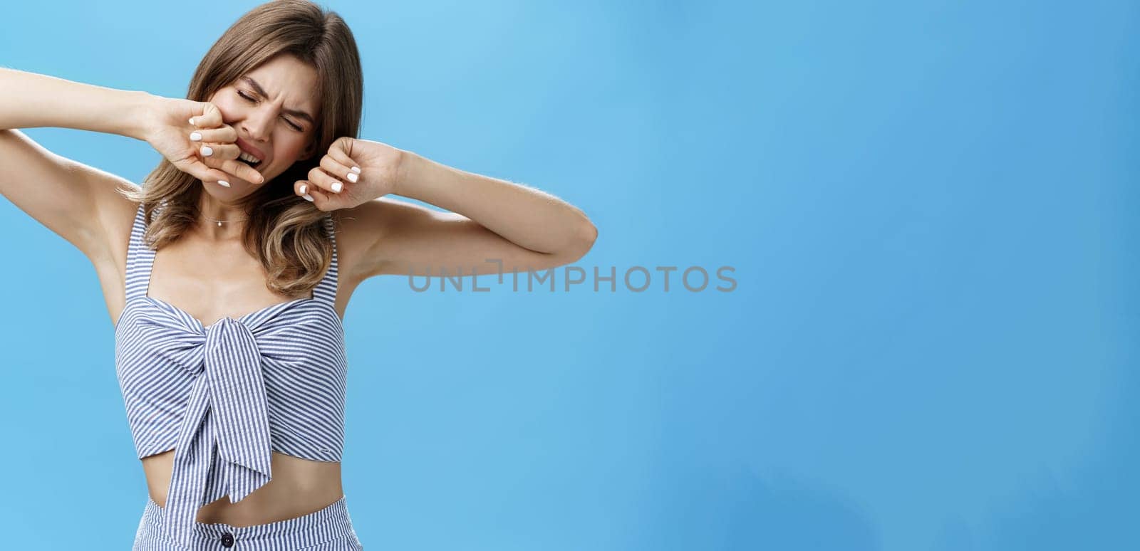 Portrait of cute sleepy european female with small tattoo on arm stretching after having nap yawning, covering open mouth with hand, standing with closed eyes after waking up over blue background. Copy space