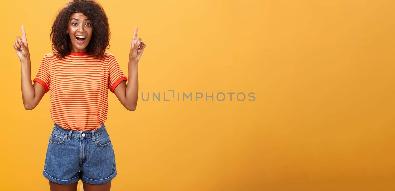 Charmed and impressed african american stylish woman seeing incredible and awesome item on sale raising hands and pointing up with broad grin being excited of new purchase over orange wall. Emotions and advertisement concept