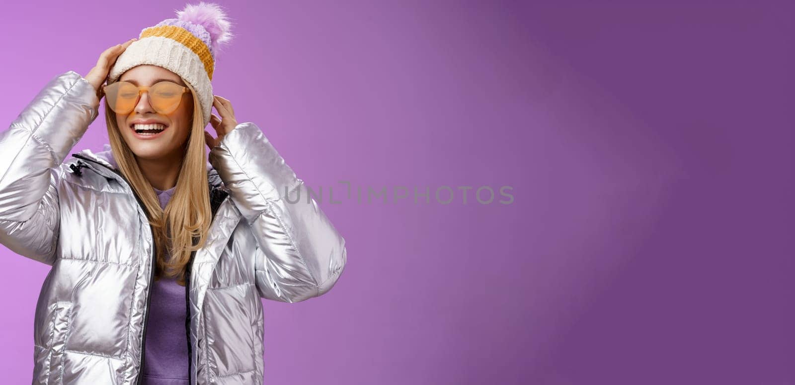 Carefree amused charming blond girlfriend having fun enjoying awesome sunny winter day ski resort vacation wearing sunglasses silver stylish jacket put on hat grinning joyfully, purple background.