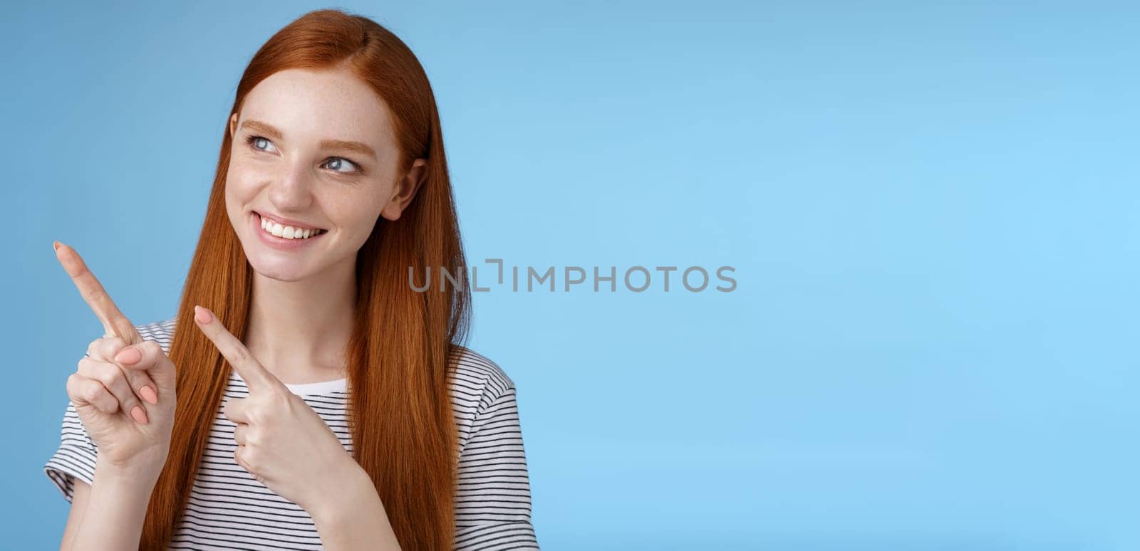 Attractive redhead female model pure clean skin ginger long hair pointing looking left intrigued smiling satisfied indicating interesting advertisement intrigued using new product, blue background.