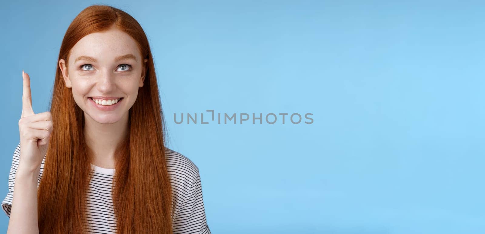 Intrigued lively happy smiling redhead european girl 20s look pointing up index fingers amused check out interesting sale holiday promo offer standing intrigued thrilled blue background by Benzoix