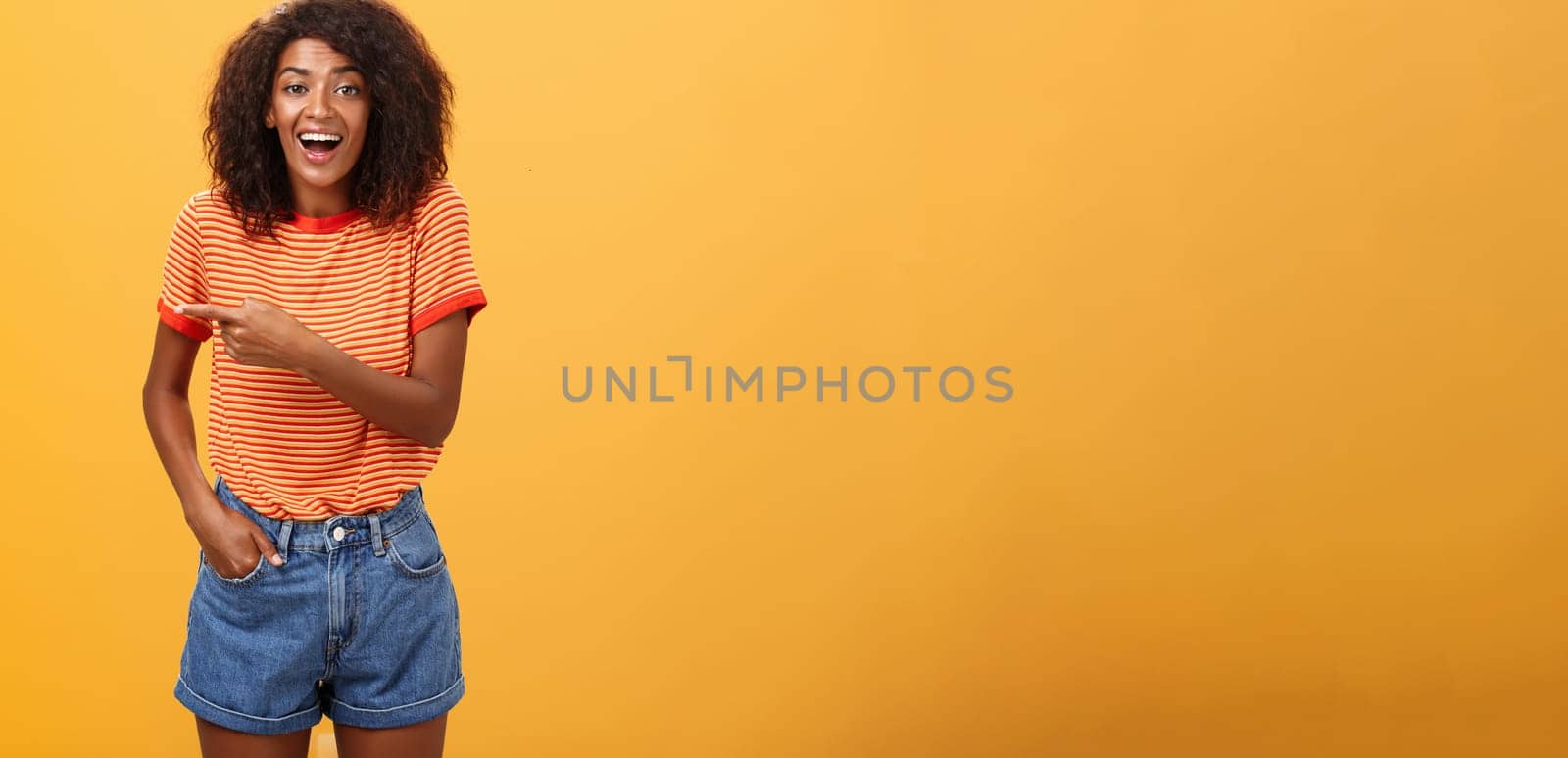 Woman laughing over funny puppy. Portrait of amused and entertained attractive stylish african-american female in casual denim shorts pointing left talking about awesome copy space over orange wall.