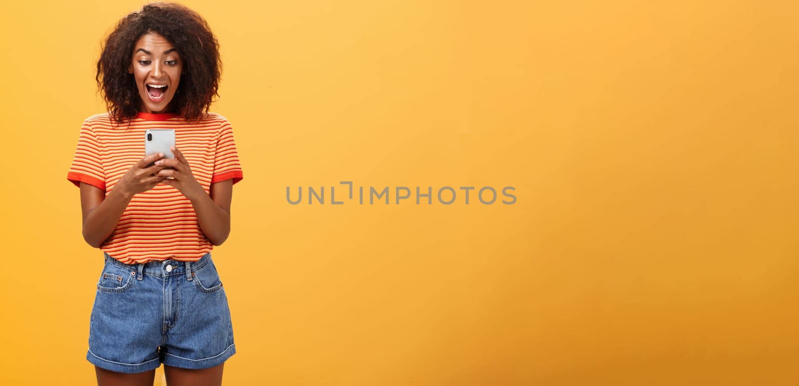 Girl expressing excitement and joy receiving awesome invitation via messages yelling from delight and happiness looking at smartphone screen impressed and thrilled over orange background.