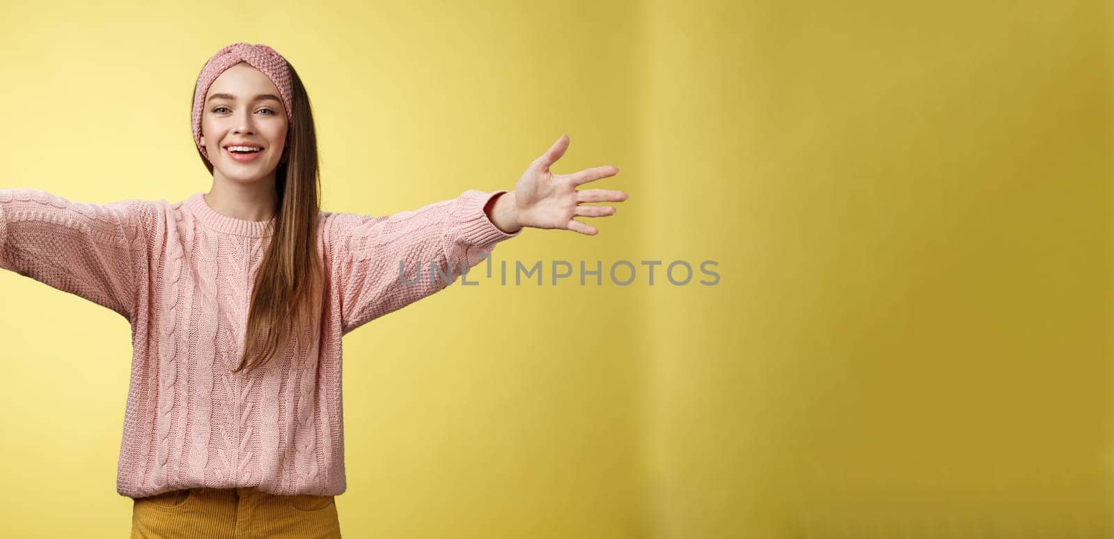 Attractive friendly positive young european woman wearing sweater, headband welcoming extanding arms and smiling at camera giving hugs, embraces friends, cuddling against yellow background. Copy space