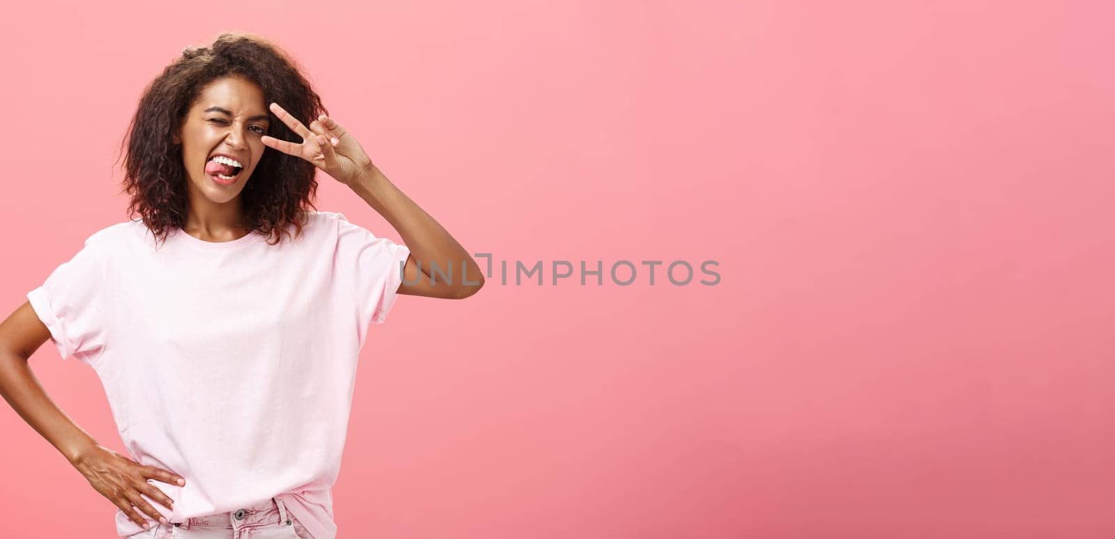 Not afraid express myself. Joyful charismatic african american woman in t-shirt with afro haircut showing tongue playfully and daring making peace sign over eye and winking posing over pink background.