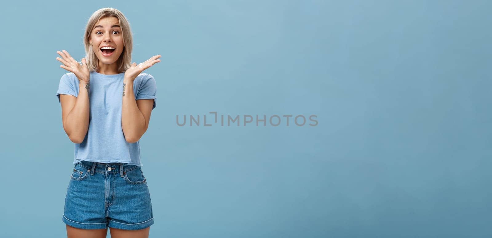 Thrilled enthusiastic happy woman in trendy t-shirt and shorts clapping hands from amazement smiling broadly and gazing with admiration at camera being surprised and joyful over blue wall. Copy space