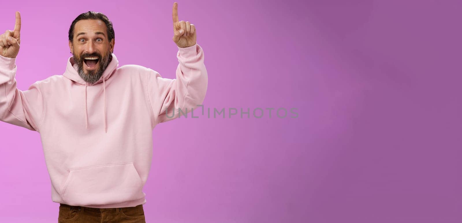 Energized happy good-looking trendy mature dad beard grey hair in pink stylish hoodie wearing son outfit pointing up index fingers thrilled favorite music group in town, standing purple background.