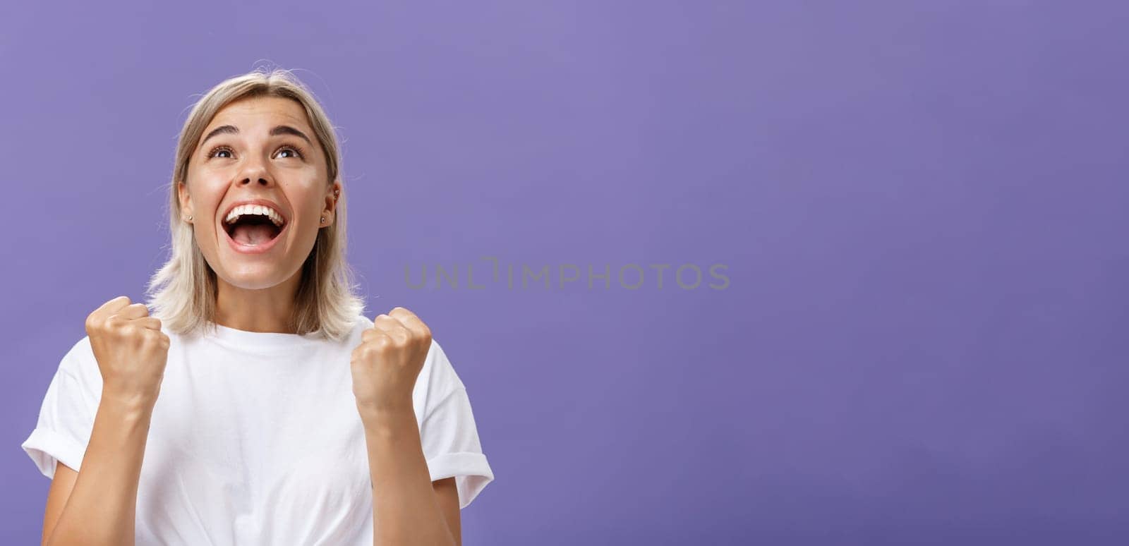 Waist-up shot of grateful delighted lucky girl with attractive tan in white t-shirt clenching fist from positive emotions gazing delighted and happy up thankful giving thanks god for fulfilling dream. Emotions concept