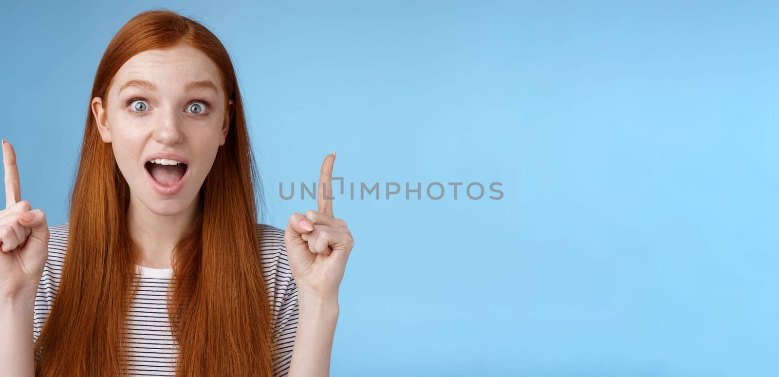 Surprised happy enthusiastic young redhead woman reacting impressed upper hanging promo pointing up index fingers drop jaw amused look thrilled camera telling about awesome offer, blue background.