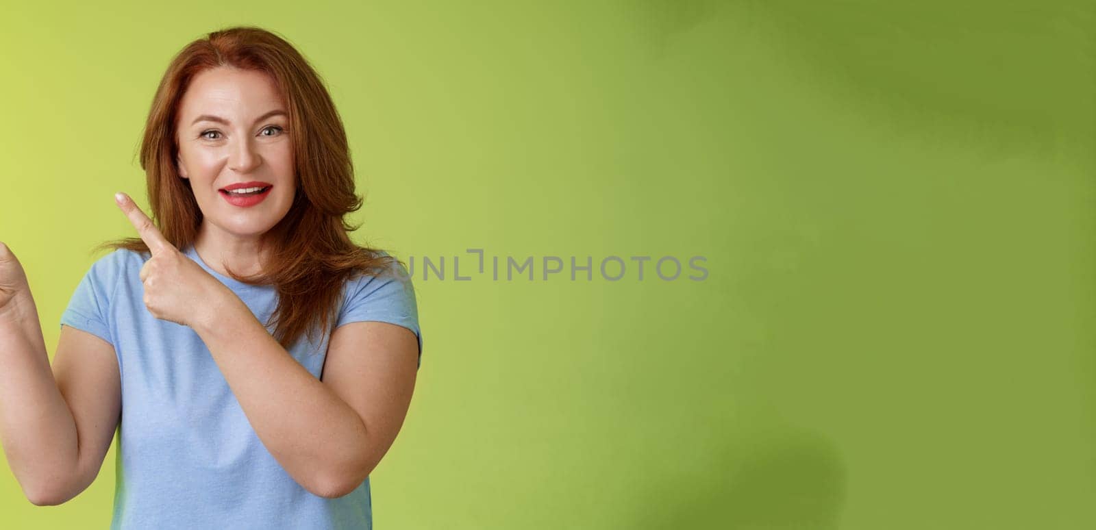 Curious enthusiastic gorgeous redhead female intrigued pointing upper left corner asking question interesting product talk shop assistant consulting promo stand green background blue t-shirt by Benzoix