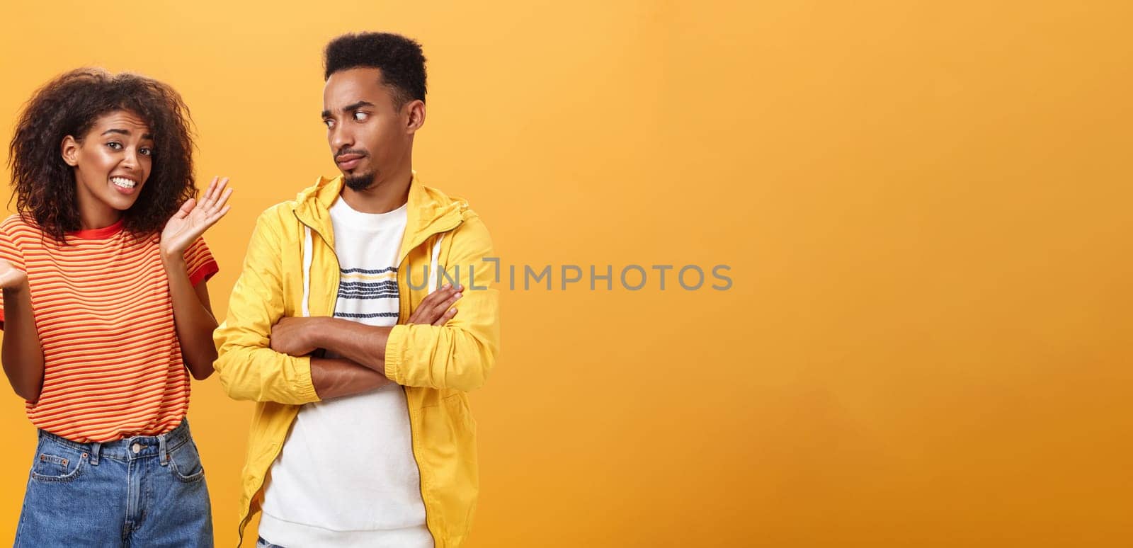 Guy thinks his friend weirdo making dumb thinks looking at cute girl with suspicious look crossing arms on chest raising eyebrow questioned while girlfriend saying sorry shrugging over orange wall. Lifestyle.