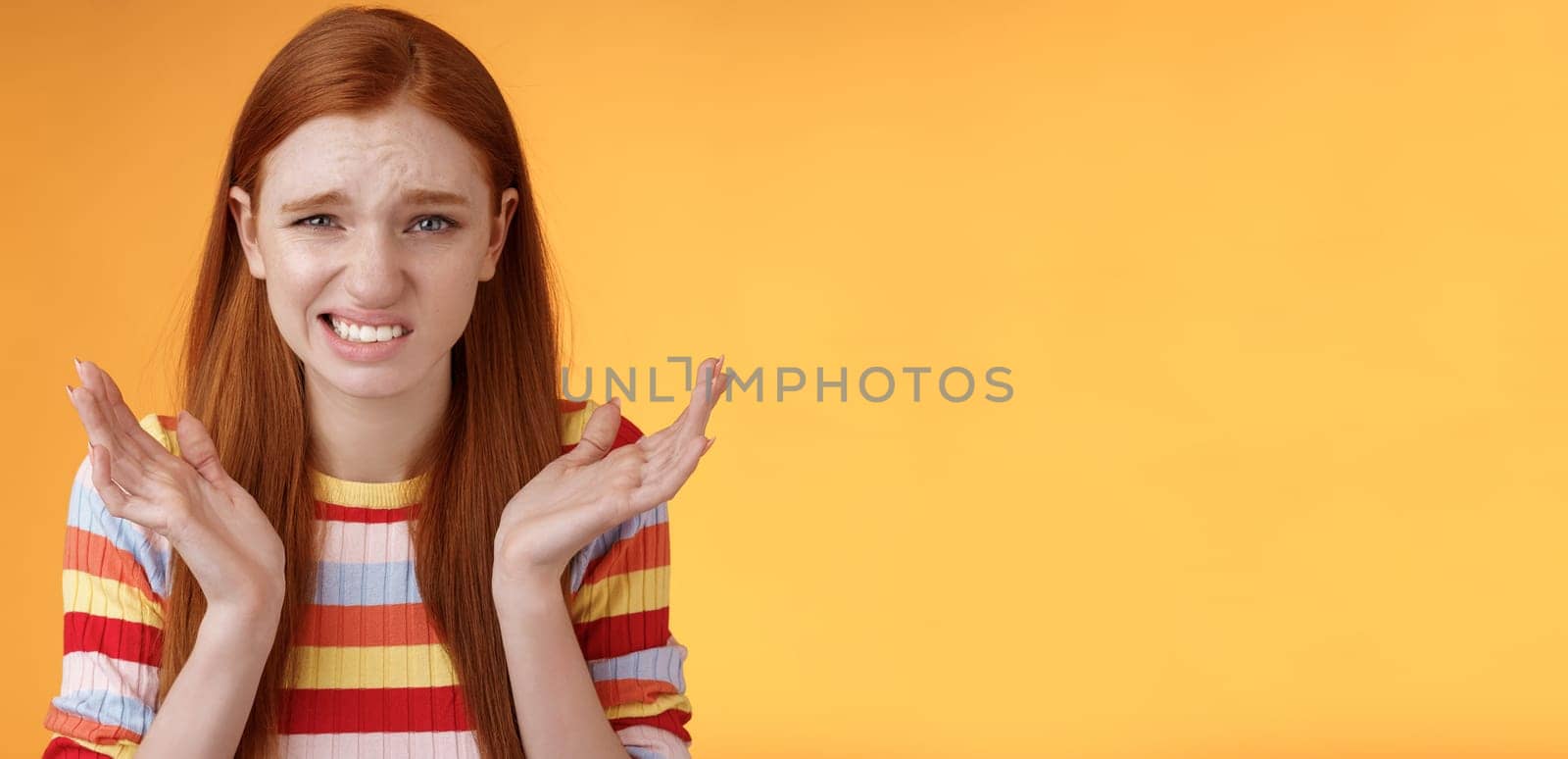 Awkward unhappy worried young redhead girl cringe feel sorry apologizing smirking smiling nervously frowning squinting spread hands sideways shrugging confused, standing orange background by Benzoix
