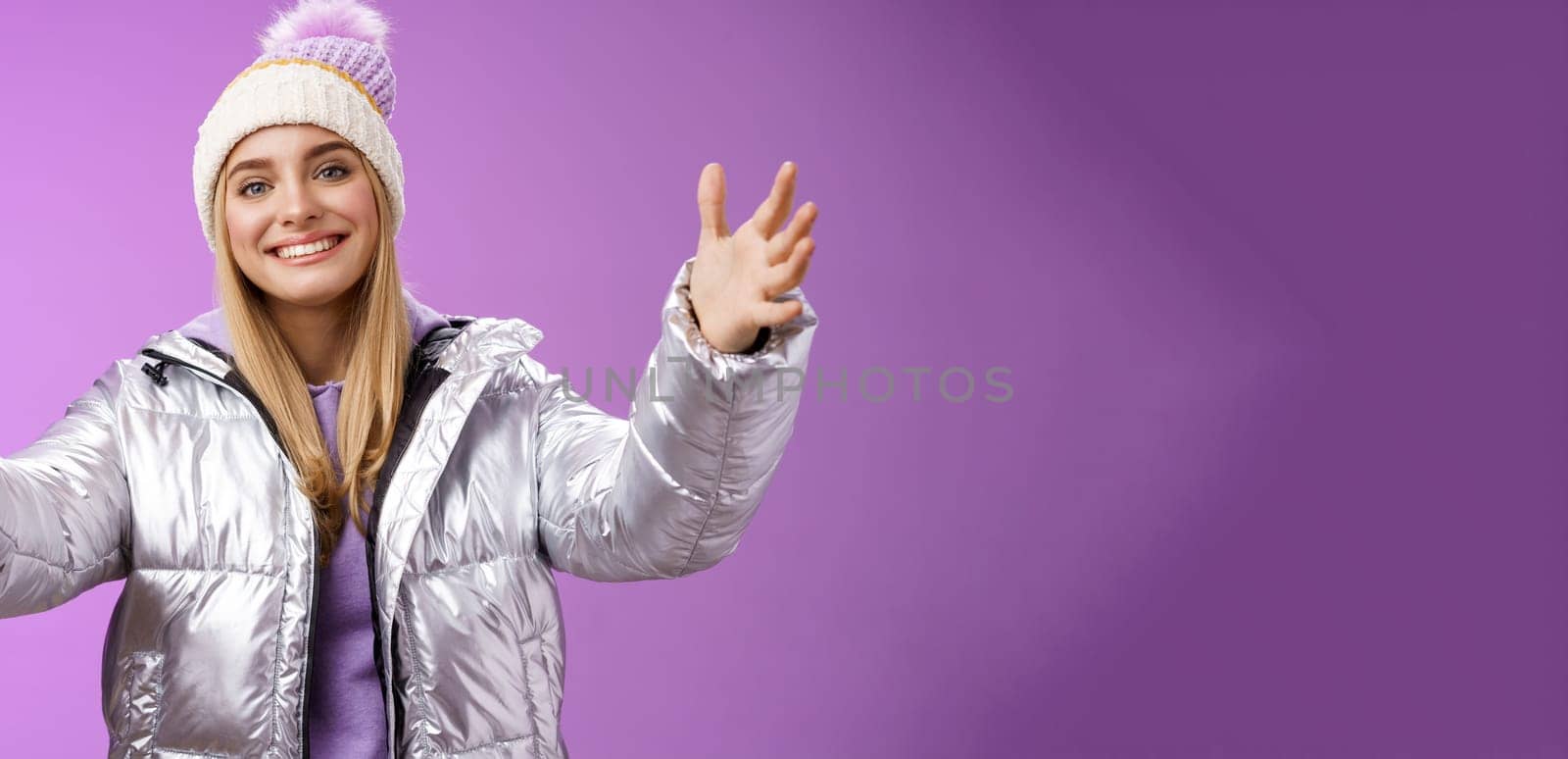 Friendly charming smiling european girl vacation wearing silver warm glittering jacket hat extend arms waiting girlfriend fall arms embrace loving person standing happily purple background.