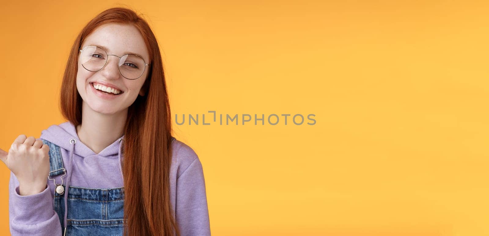 Cute helpful friendly-looking joyful european redhead woman show thumb left smiling delighted laughing pointing where find awesome store telling about interesting new promo offer, orange background.