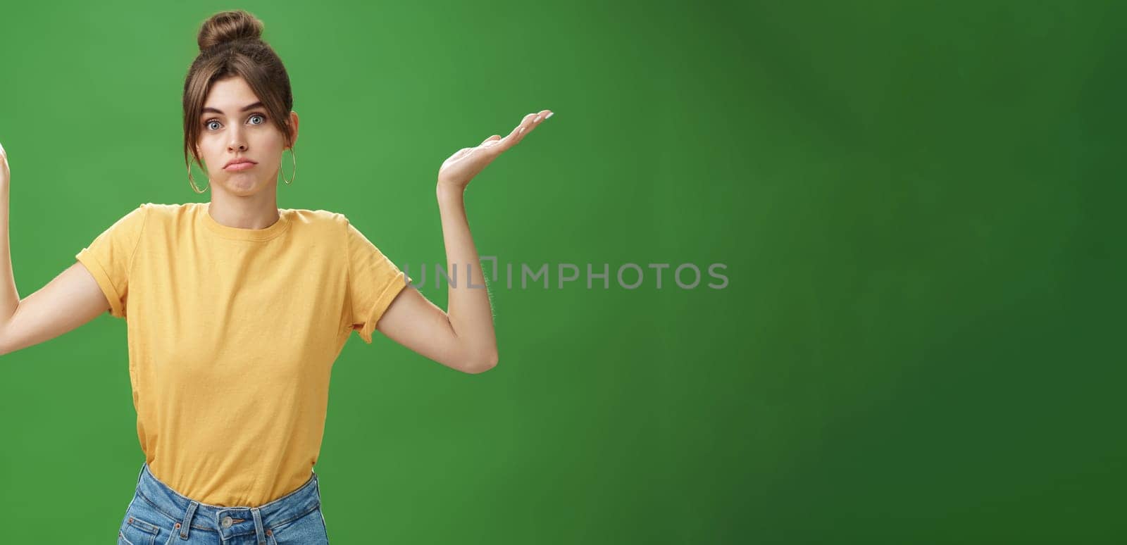 Girl do not know anything. Portrait of confused unsure cute woman with combed hair shrugging with raised hands pouting looking clueless at camera standing questioned against green background. Body language concept