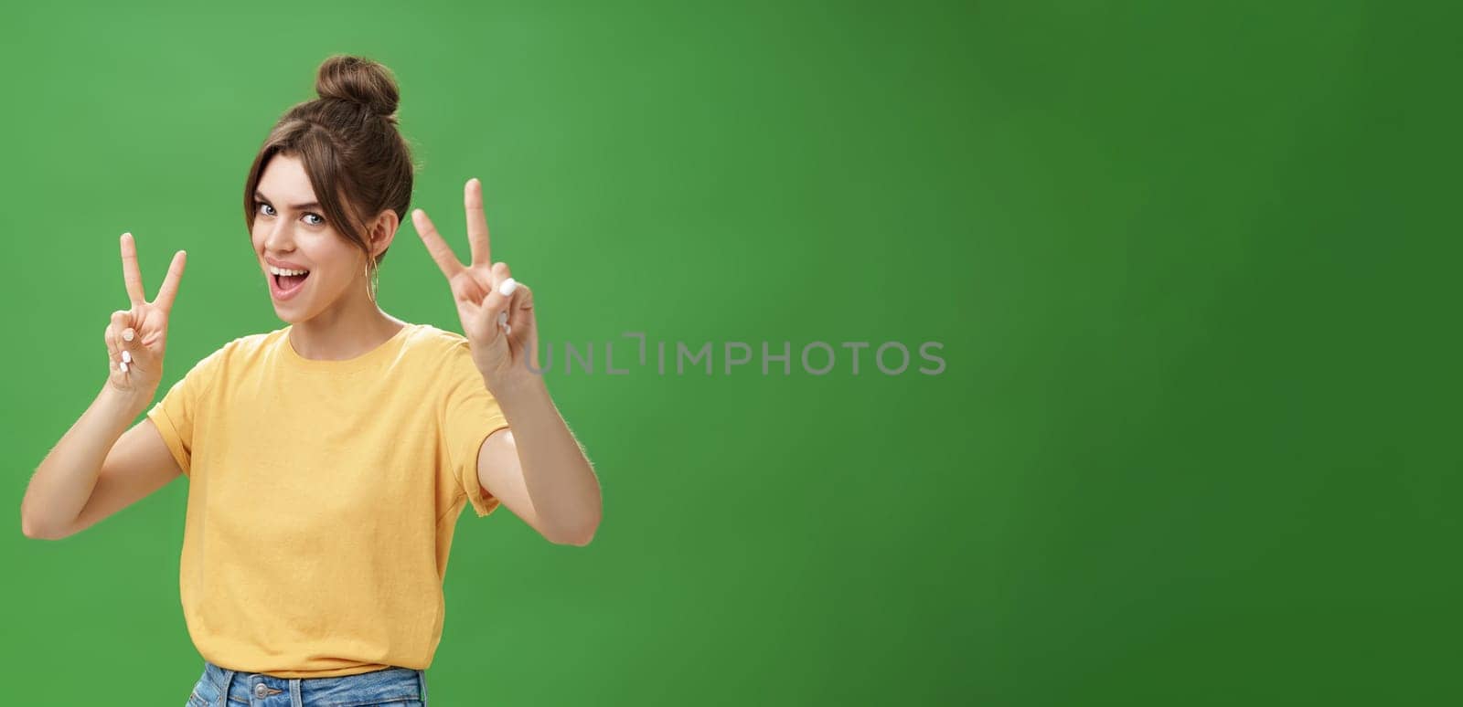 Indoor shot of enthusiastic excited and happy daring girl with combed hair tattoo and cute diasdema showing peace signs bending backwards standing in cool energized pose over green background by Benzoix