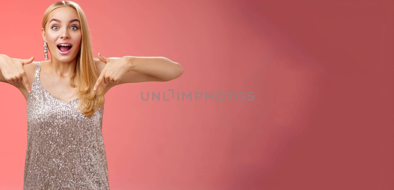 Impressed excited attractive glamour blond girl in silver glittering dress gasping thrilled pointing down glance camera fascinated check out fabulous awesome promo, standing amused red background.