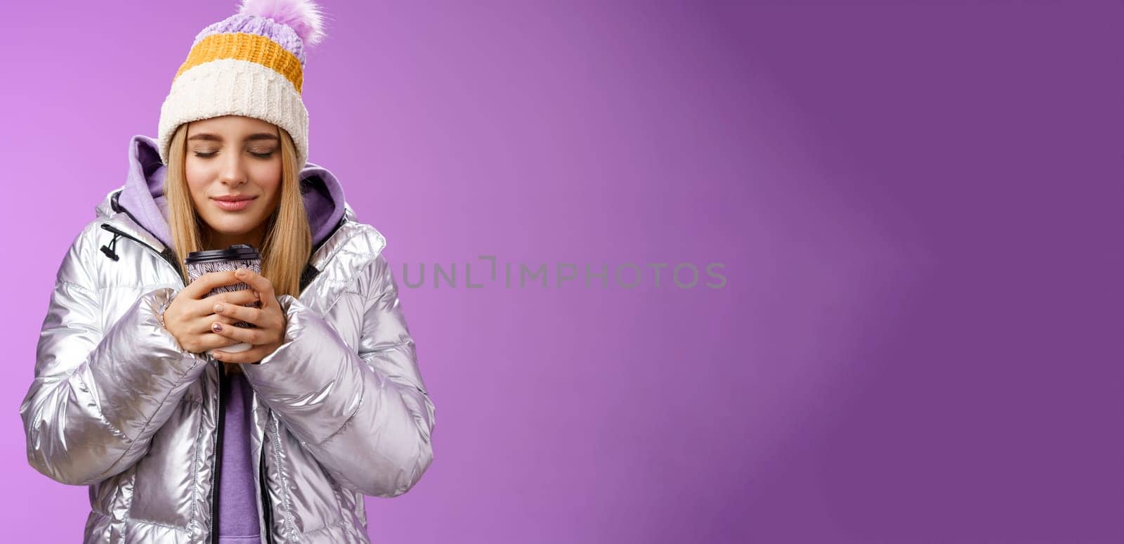 Charming tender romantic blond girl sniffing nice smell warm tasty coffee holding take-away cup warm hands close eyes look delighted wearing winter jacket hat enjoy vacation, purple background by Benzoix