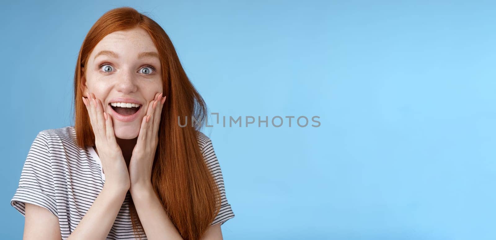 Amused happy dreamy young excited redhead female fan looking fascinated camera smiling admiration delighted touch face thrilled express emotional cheerful surprise, blue background by Benzoix