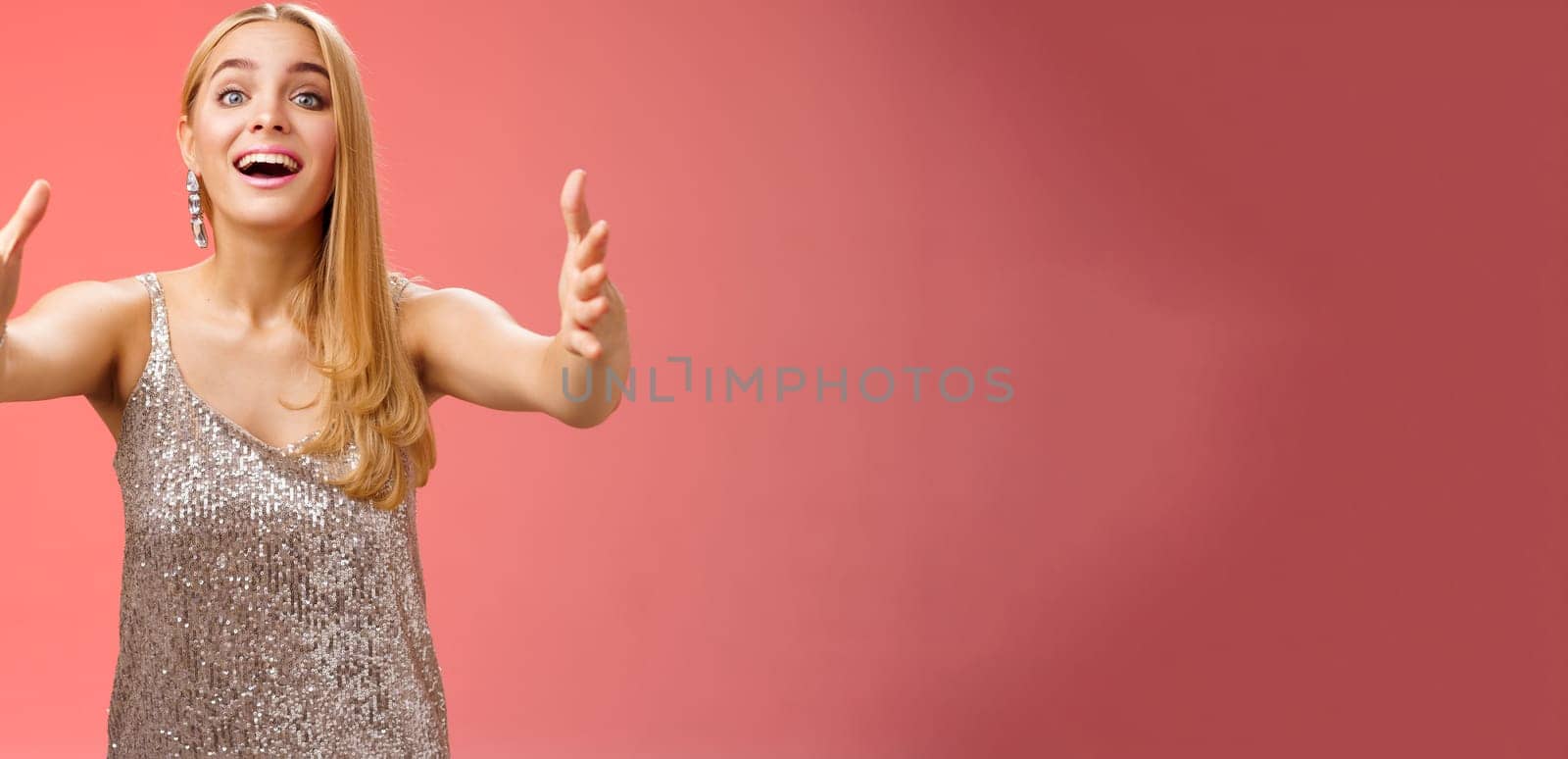 Excited charmign touched hearwarming young blond woman in silver party glamour dress stretch hands towards camera amused wanna hug cuddle hold cute puppy arms, standing red background.