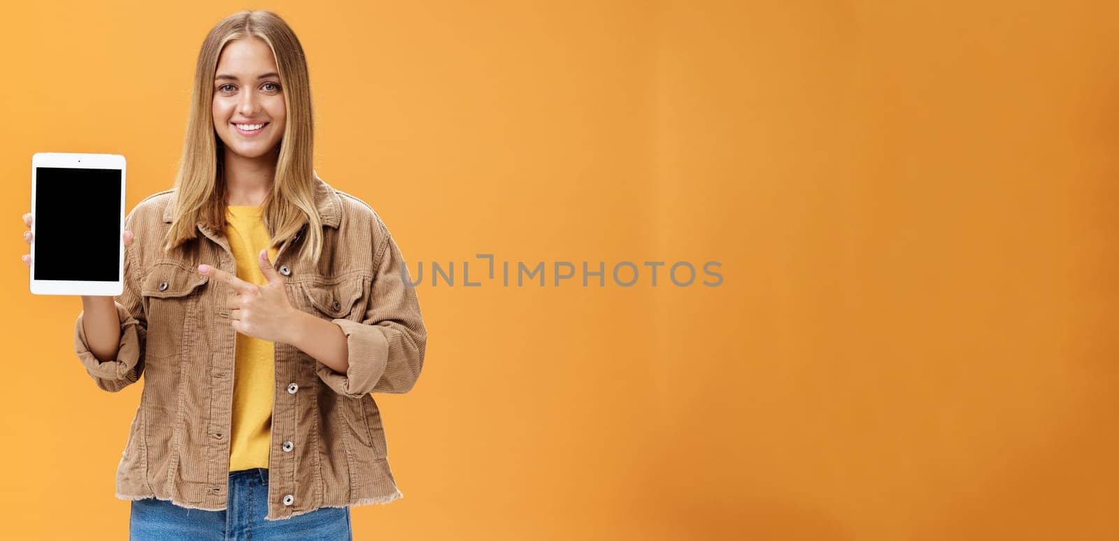 Get ready for autumn semister with new digital tablet. Portrait of charming female in corduroy jacket showing gadget screen pointing at device and smiling pleased and friendly over orange wall. Technology and advertising concept