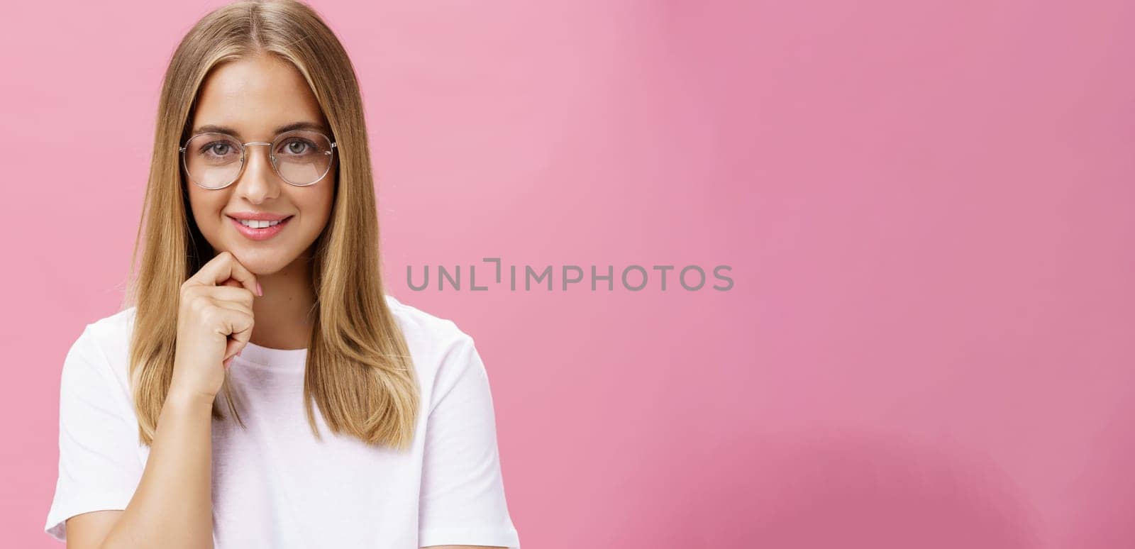 Woman listen with interest and smart look touching chin in thoughtful gesture smiling pleased, amused, looking self-assured at camera with intelligent expression over pink background, wearing glasses.