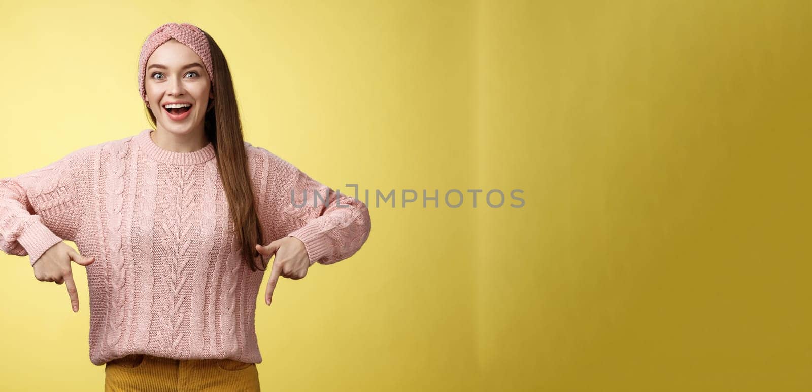 Euphoric attractive young trendy girl in knitted band wearing sweater grinning excited enthusiastic pointing down thrilled of awesome promotion standing amused and overwhelmed over yellow background by Benzoix