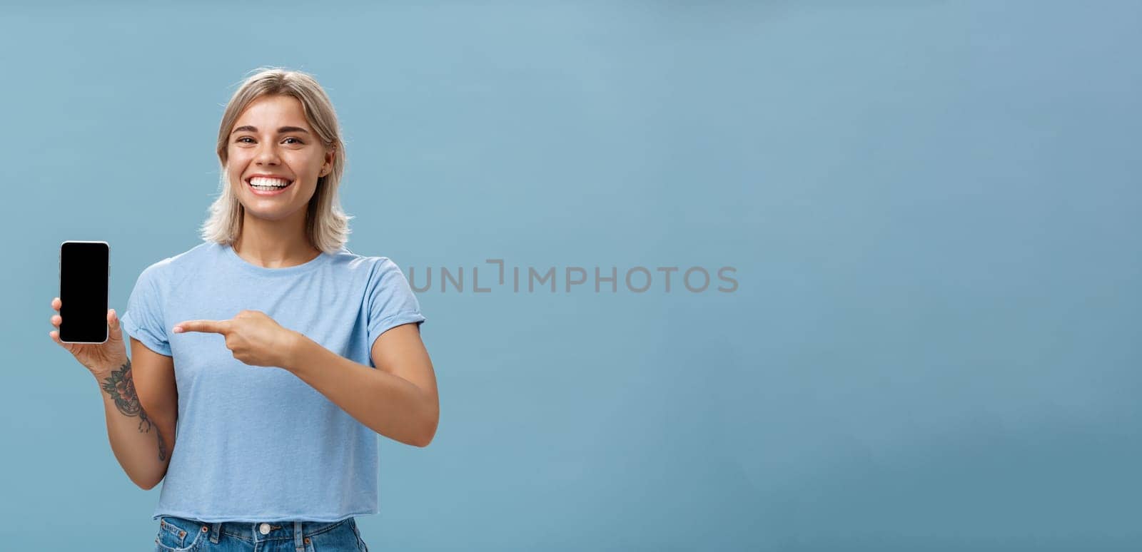 Cute enrgized girl showing smartphone. Charming blonde young female with cool tattoos laughing and smiling from happiness and joy pointing at device holding phone with screen faced to camera.