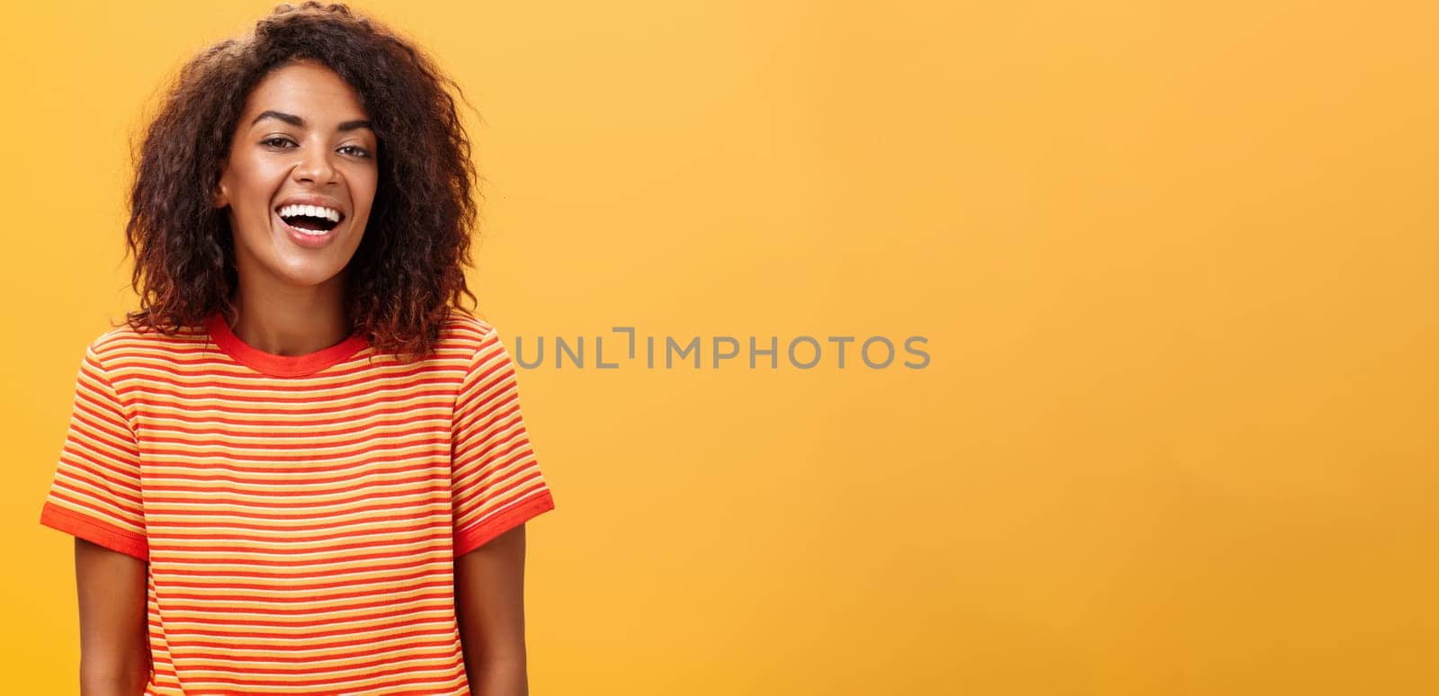 Waist-up shot of outgoing happy charming dark-skinned female with curly hairstyle laughing joyfully posing in striped trendy t-shirt over orange background enjoying nice casual conversation by Benzoix