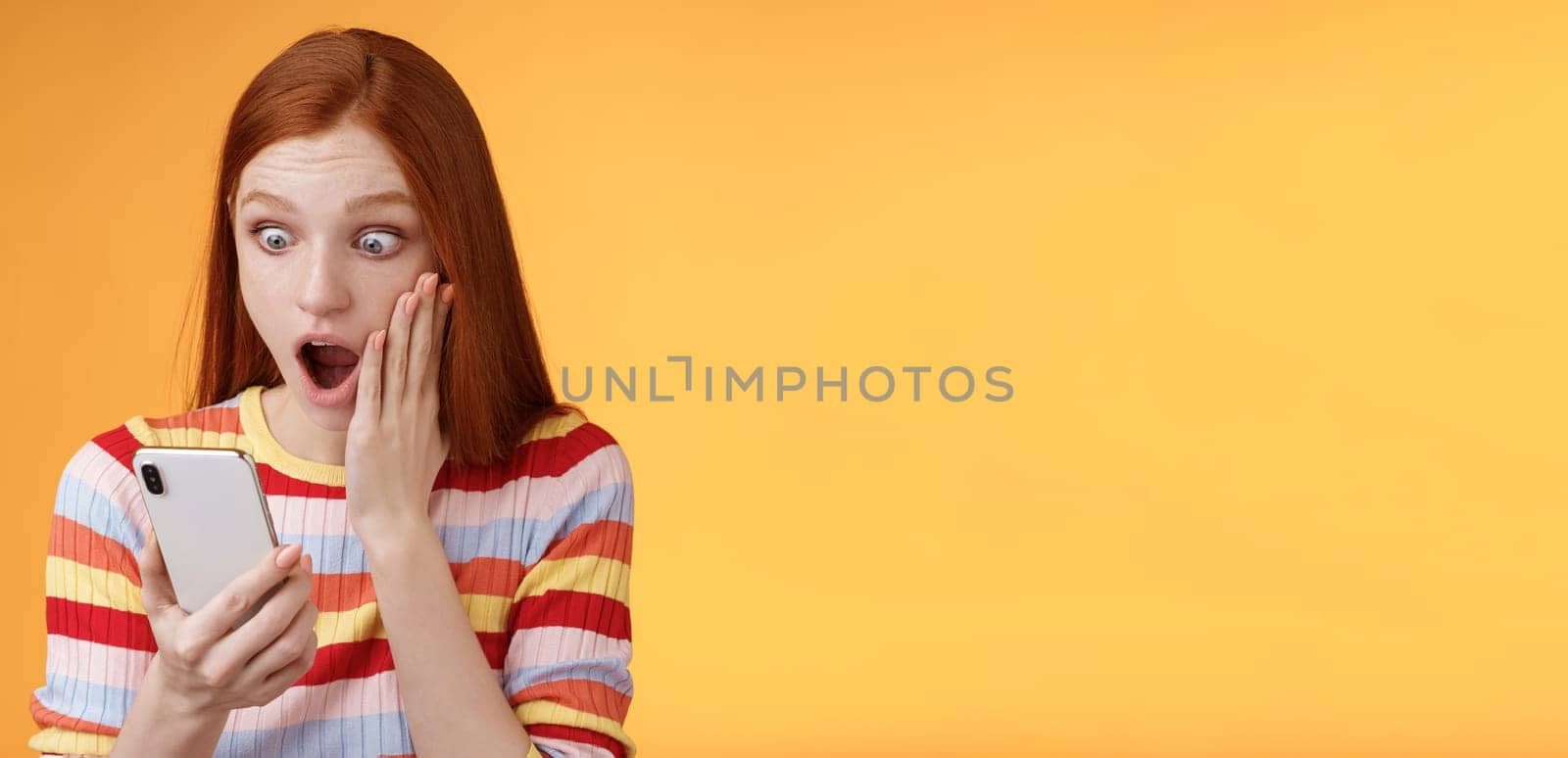 Omg what heck. Portrait shocked concerned young redhead sensitive impressed redhead woman stare smartphone display touch cheek drop jaw stunned surprised standing orange background hold phone by Benzoix