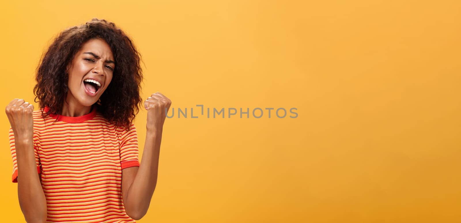 Cheerful delighted and enthusiastic african american woman with afro hairstyle clenching raised fists yelling yes from triumph and joy of success standing satisfied of victory over orange wall. Lifestyle.