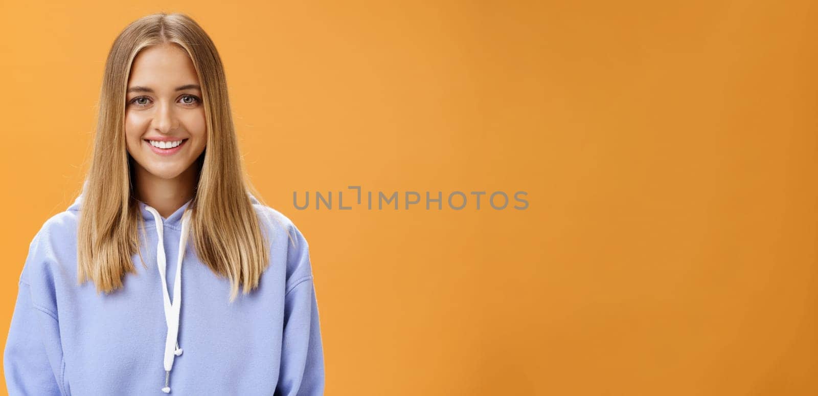Waist-up shot of pleasant kind and happy friendly-looking young fair-haired girl in comfortable hoodie smiling cheerfully at camera with delighted carefree look posing against orange background by Benzoix