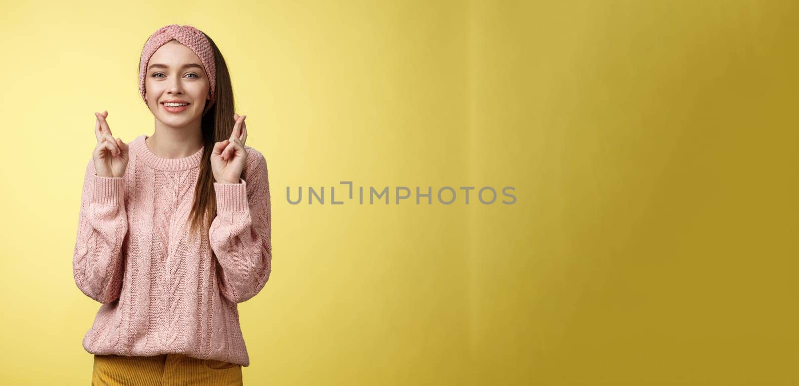 Cross my fingers for you. Supportive cute young european girlfriend wearing sweater, glamour headband making luck, wish gesture smiling hopeful having faith dream come true over yellow background by Benzoix