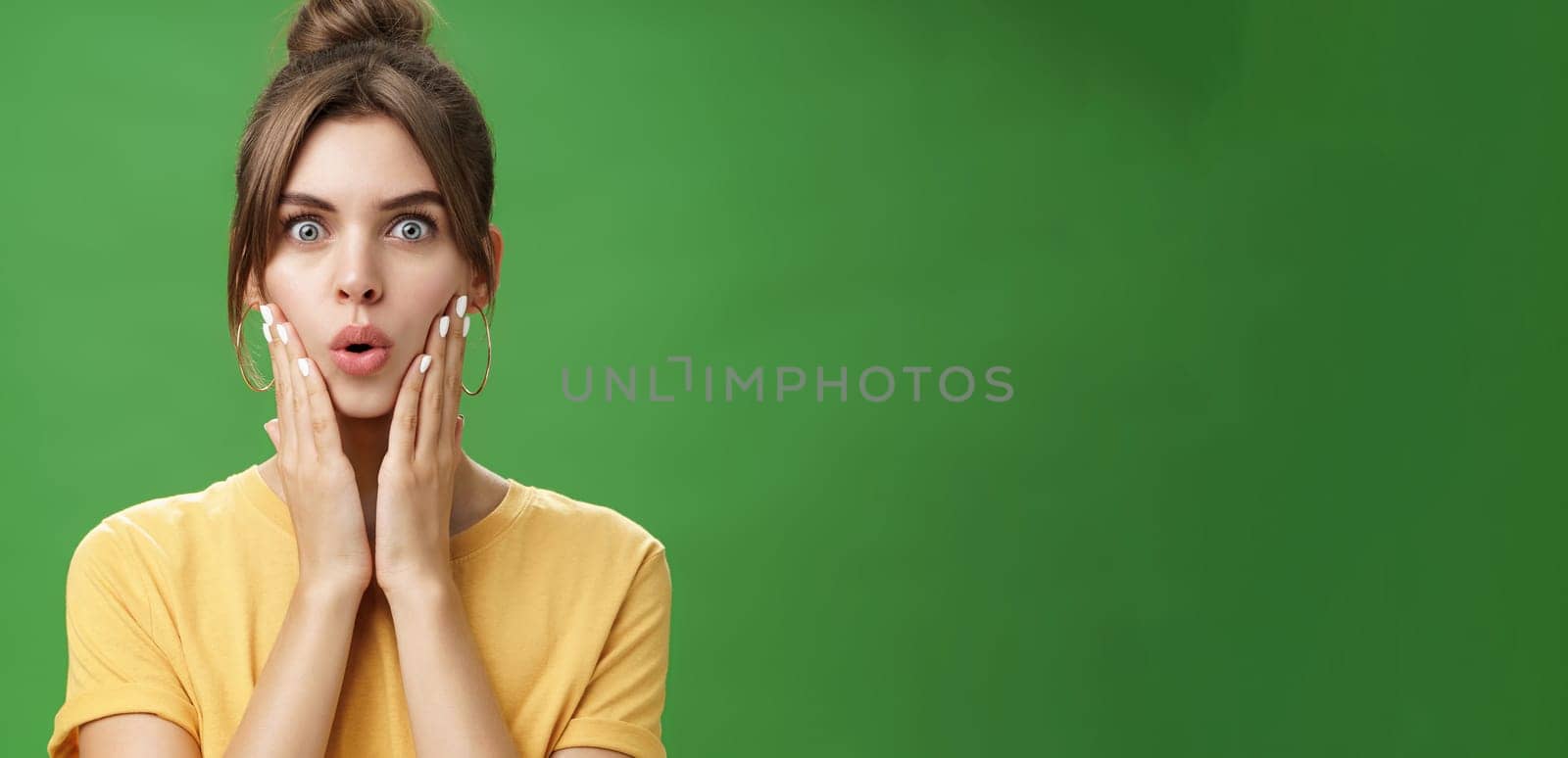 Portrait of impressed and shocked cute feminine woman in yellow t-shirt folding lips from excitement and interest touching cheeks surprised reacting to amazing rumor posing over green background. Body language concept