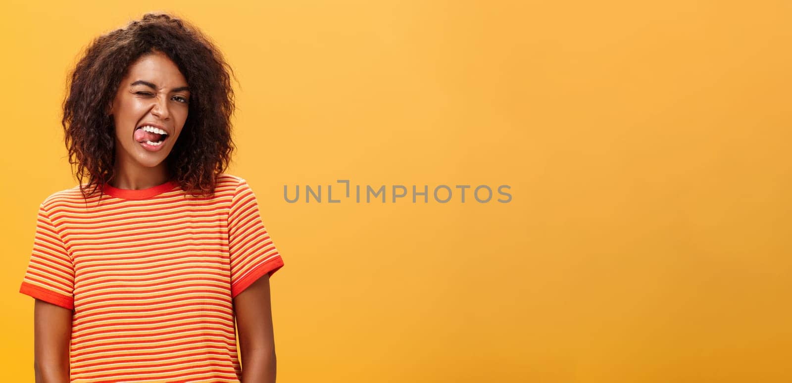 Portrait of daring and emotive confident flirty woman with afro hairstyle winking joyfully showing tongue posing carefree and enthusiastic against orange background flirting with hot guy. Copy space