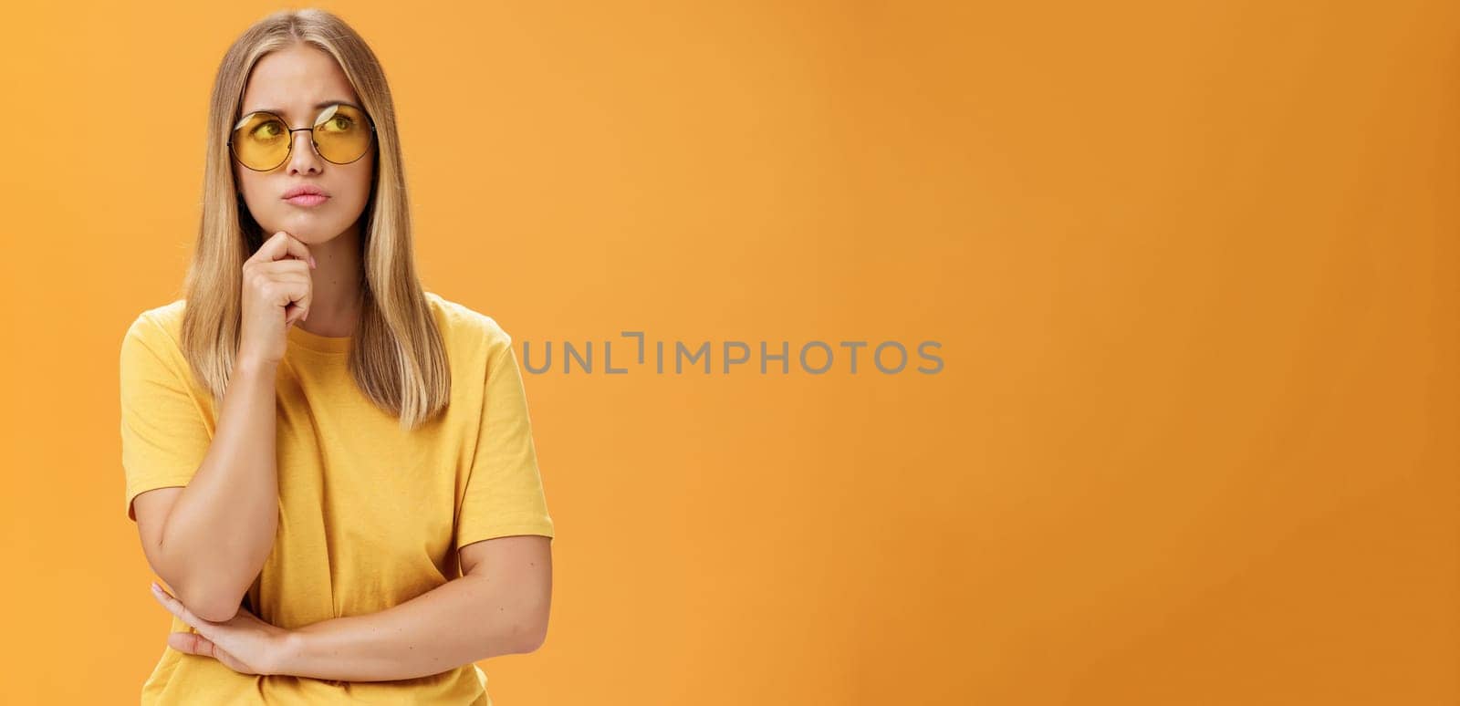 Troubled and concerned young stylish woman facing troublesome problem thinking holding hand on chin pursing lips looking at upper right corner thoughtfully standing against orange background by Benzoix