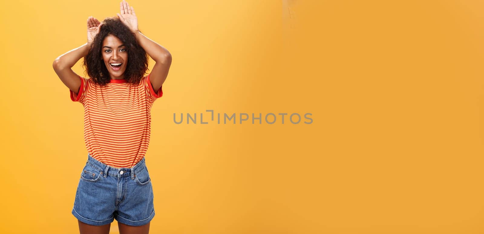 Let me be your bunny. Portrait of charming enthusiastic and charismatic happy dark-skinned female with afro hairstyle holding palms on head like animal ears making cute face over orange background.