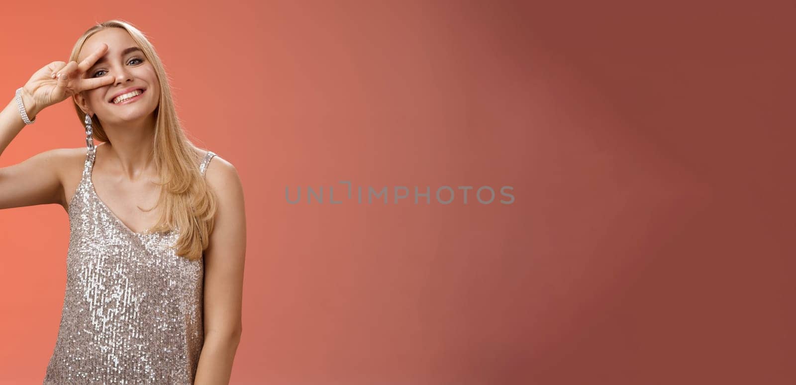 Optimistic lucky elegant european blond young 25s wife in silver glittering dress having fun enjoying celebration party dance floor showing peace victory sign eye smiling delighted, red background.