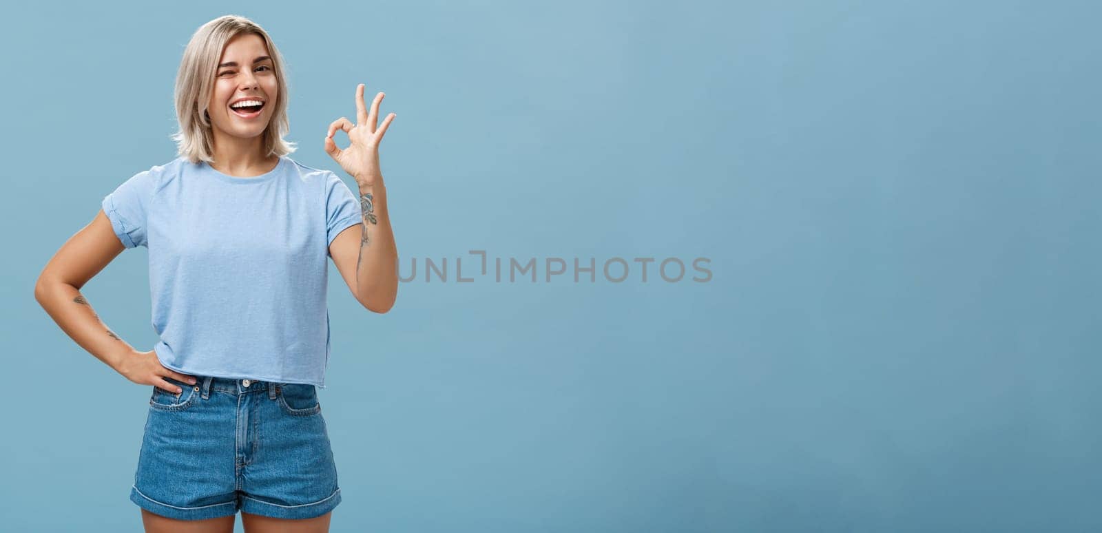 Confident and smart girl got everything under control. Portrait of optimistic good-looking girl with fair hair and tanned skin winking showing okay or perfect gesture standing over blue wall.