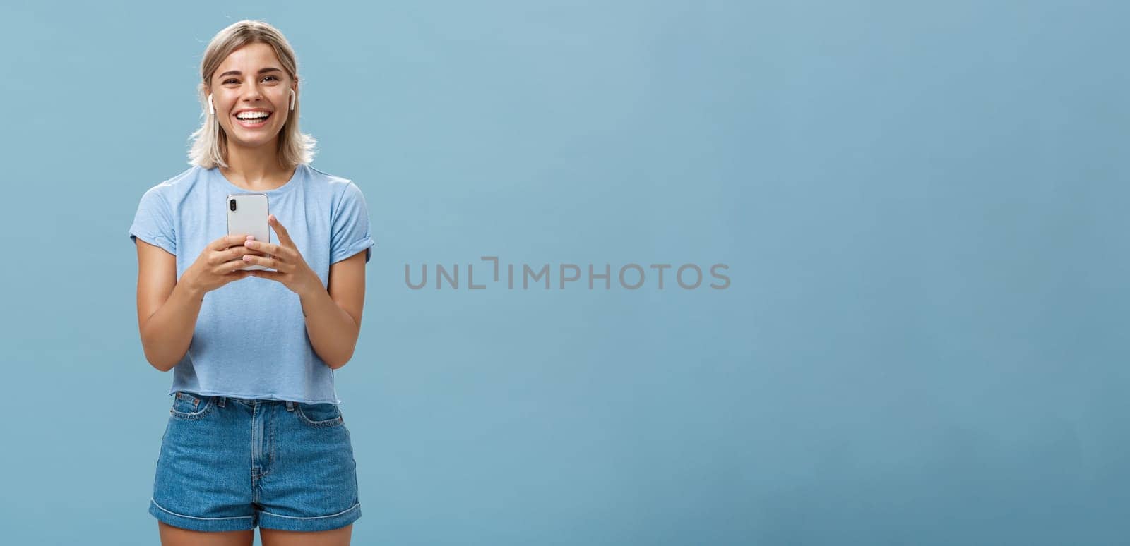 Satisfied happy and carefree young attractive blonde woman in casual outfit smiling broadly from happiness and delight listening music in wireless earphones holding smartphone over blue wall by Benzoix