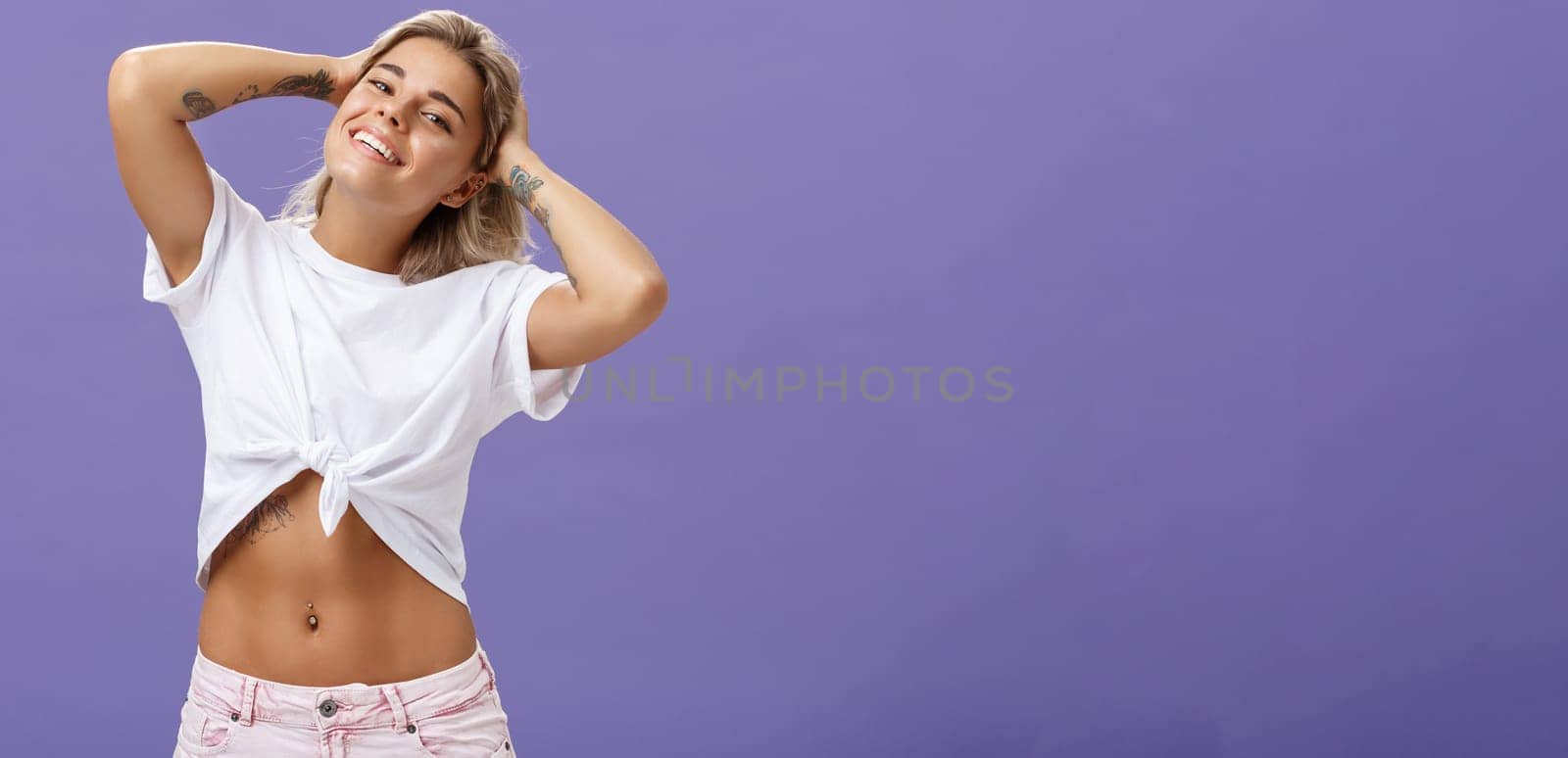 Waist-up shot of creative carefree and happy good-looking young tanned girlfriend with perfect body and tattoos holding hands behind head smiling joyfully, having great time over purple wall. Emotions concept