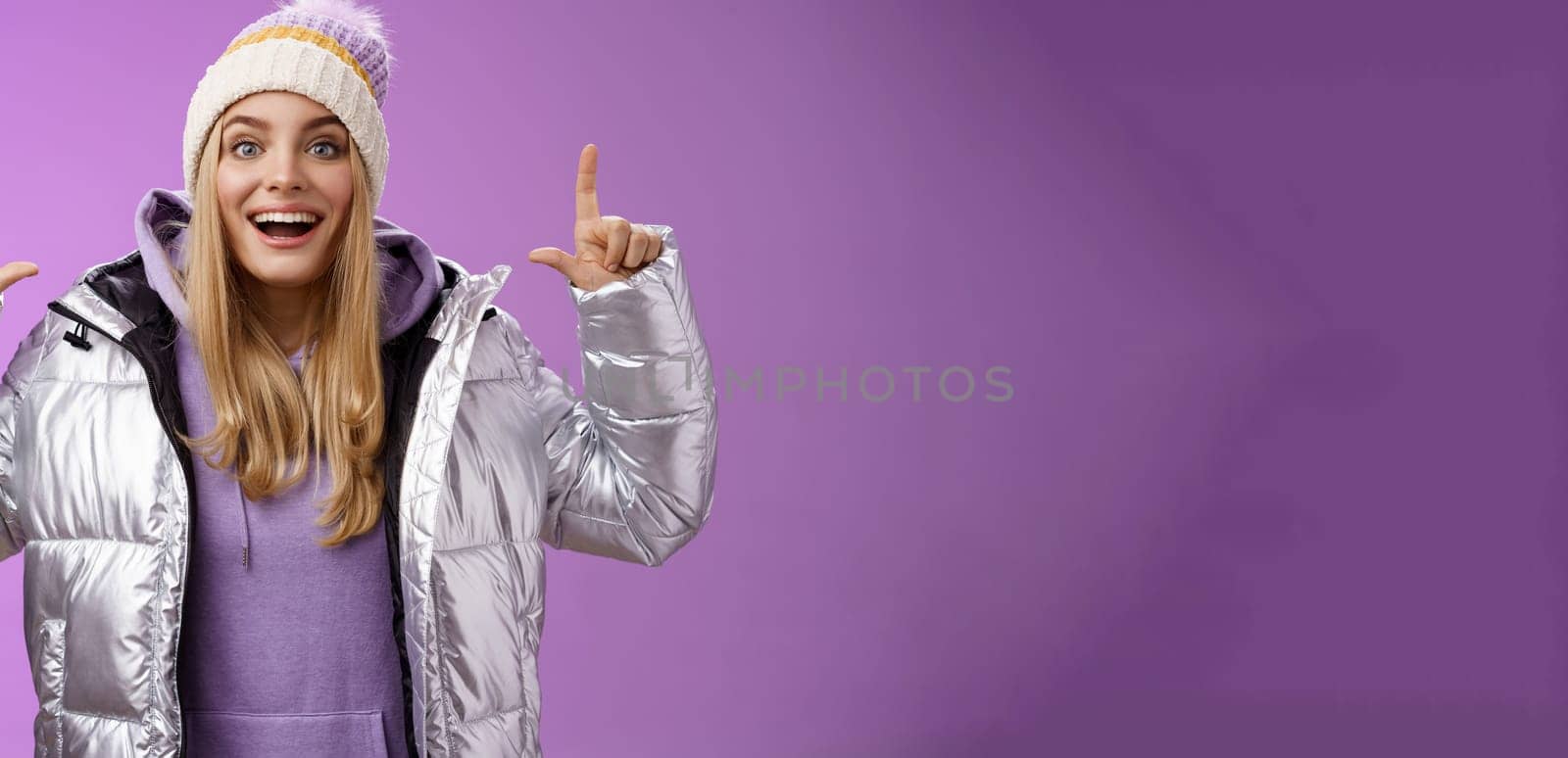 Lifestyle. Excited carefree cheerful fair-haired european girl in silver jacket winter hat raising hands pointing up have excellent idea smiling broadly speaking passionately standing purple background.