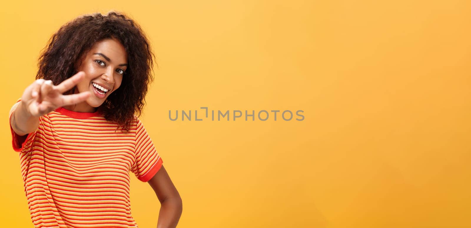 Hey peace my friend. Charming outgoing and confident carefree dark-skinned girl with afro hairstyle in trendy t-shirt pulling hand with victory gesture towards camera smiling over orange wall.