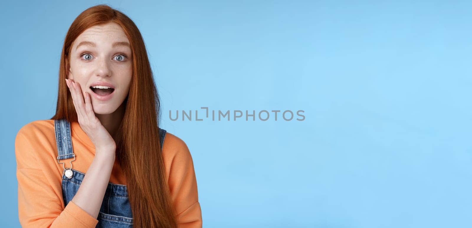 Amazed friendly sensitive impressed redhead girl learn awesome pleasant news open mouth wondered astonished smiling glad friend touch cheek amused, standing blue background happy. Copy space