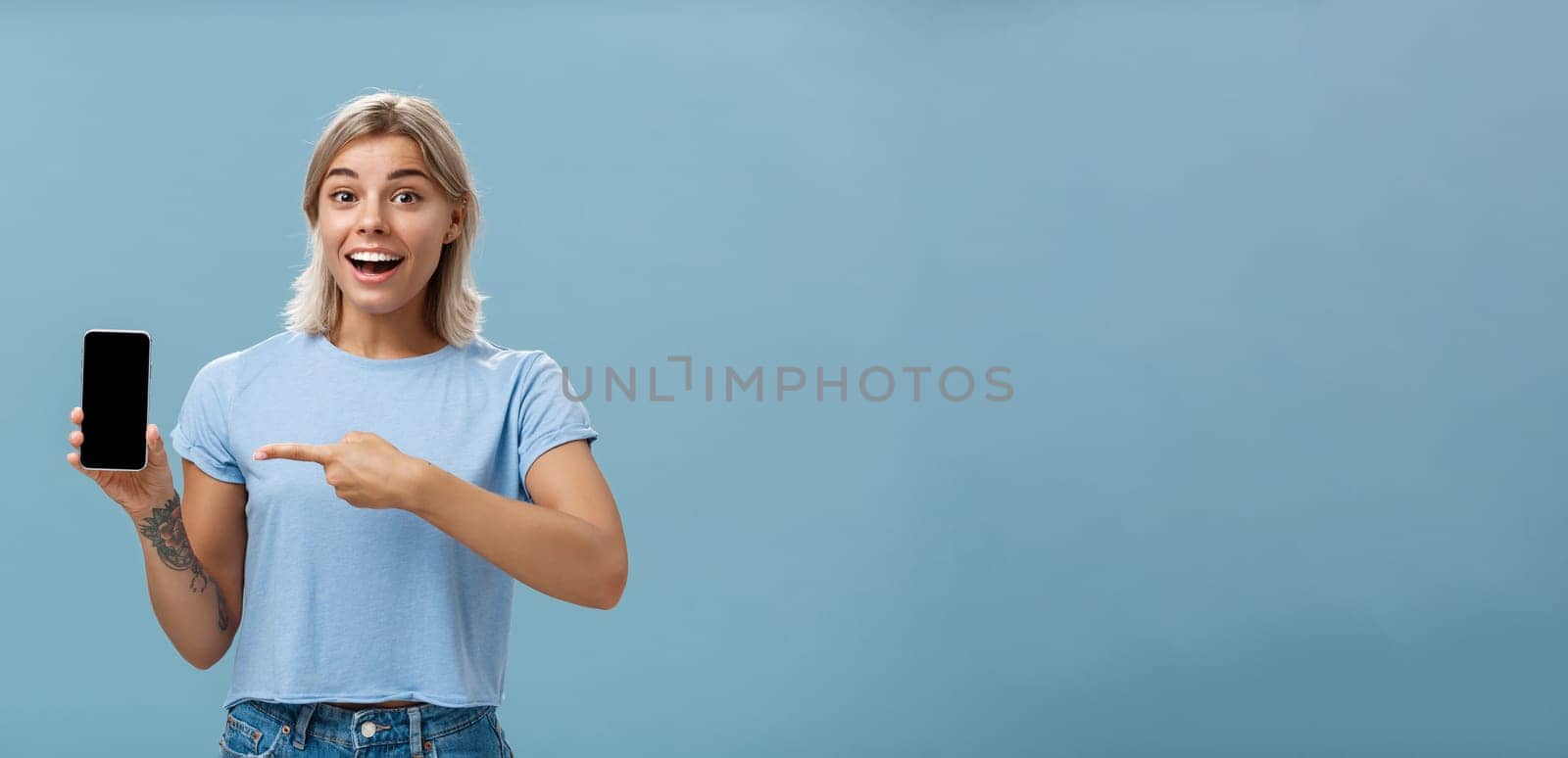 Lifestyle. Waist-up shot of thrilled and impressed good-looking female student in casual t-shirt smiling joyfully pointing at smartphone screen showing awesome place via internet to friend over blue background.