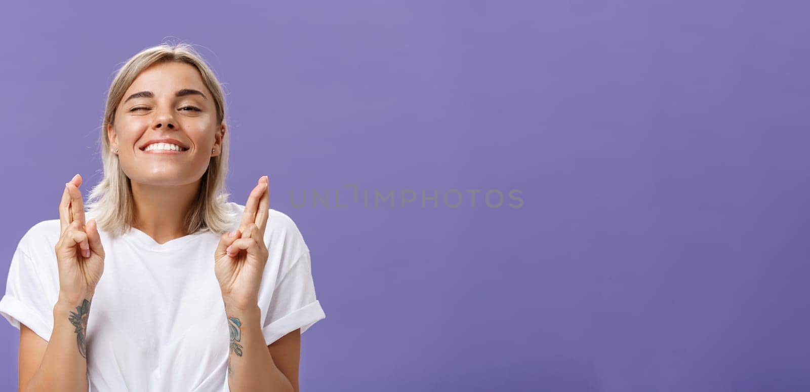 Waist-up shot of hopeful optimistic attractive stylish woman in white t-shirt with tattooed arms winking smiling joyfully while crossing fingers for good luck making wish over purple background. Body language concept