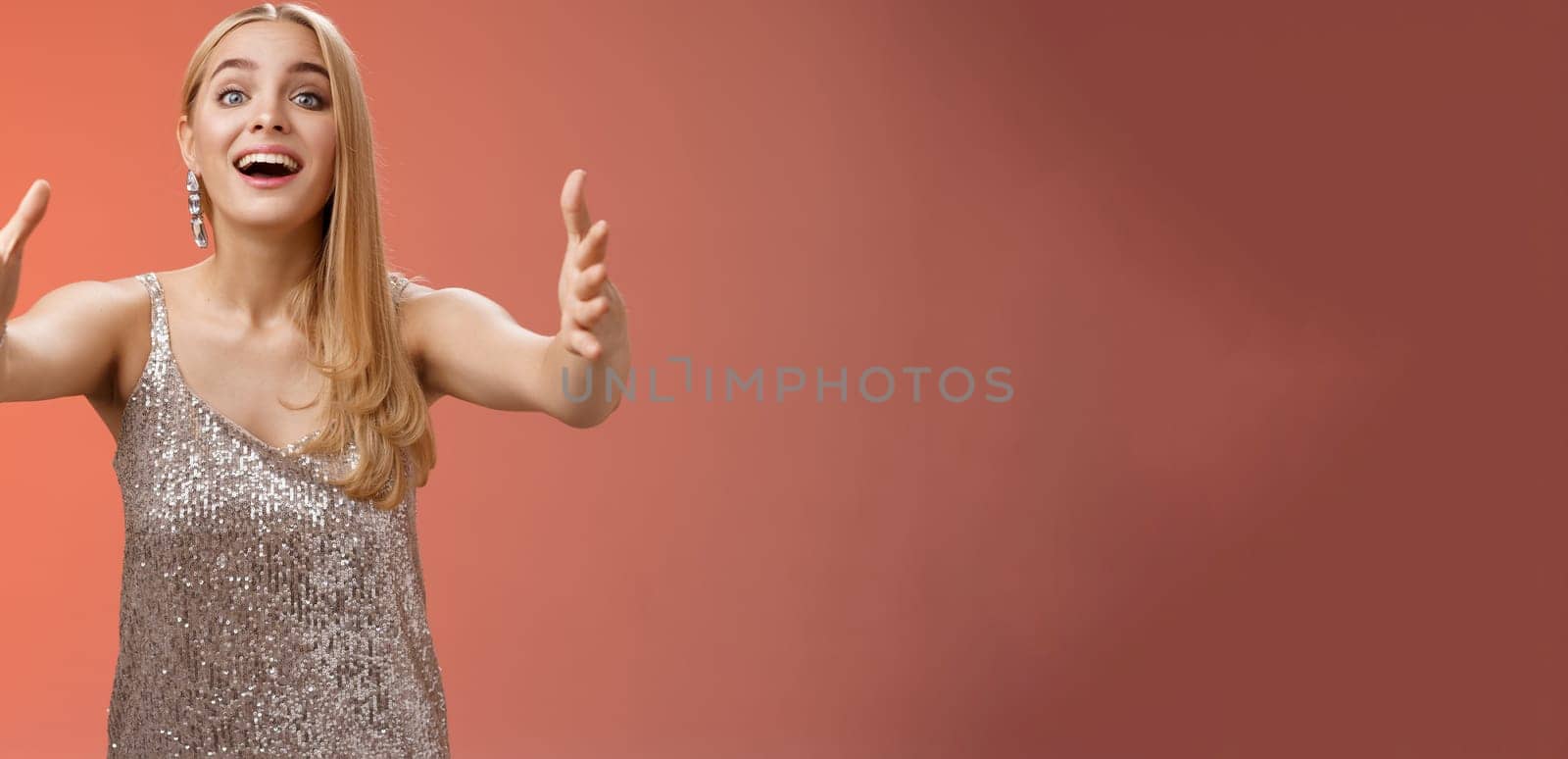 Excited charmign touched hearwarming young blond woman in silver party glamour dress stretch hands towards camera amused wanna hug cuddle hold cute puppy arms, standing red background.