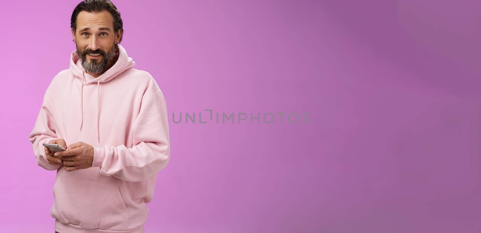 Stylish handsome adult bearded guy 40s in trendy pink hoodie holding smartphone texting look camera unbothered cool confident distracted playing awesome new app game, purple background.